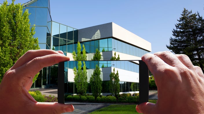 Hands holding a smartphone to capture a photo of a modern office building for photo-verified service documentation.